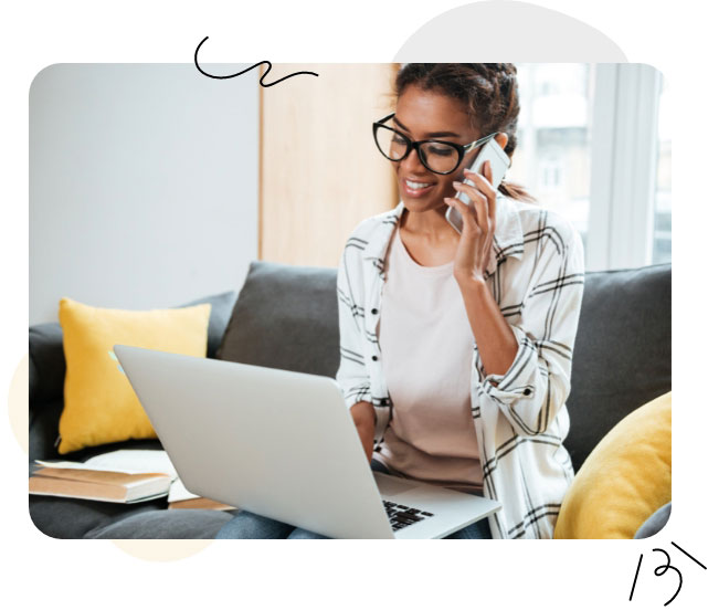 Une femme portant des lunettes est assise sur un canapé, tenant un ordinateur portable et un téléphone dans ses mains, effectuant plusieurs tâches à la fois.