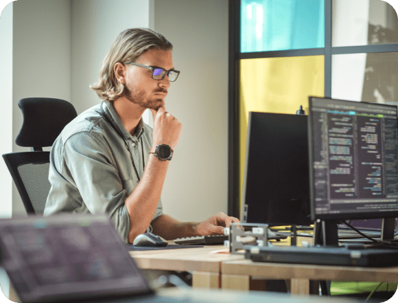 A man wearing glasses sits at a desk, working on two computers side by side.
